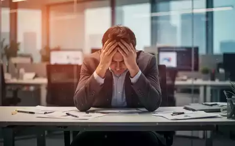 Eine Person sitzt an einem modernen Schreibtisch, den Kopf in den Händen, umgeben von verschwommenen Büroeinrichtungen. Die Atmosphäre ist von Erschöpfung und Stress geprägt. - Foto: über boerse-global.de