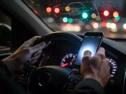Nahaufnahme einer Hand, die ein Smartphone am Lenkrad hält, mit verschwommenem Straßenverkehr im Hintergrund. - Foto: über boerse-global.de