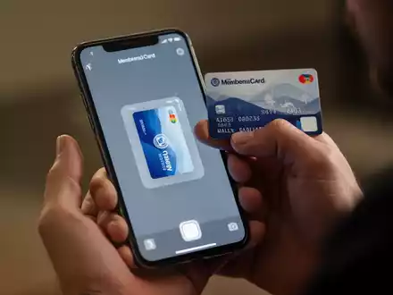 Eine Hand hält ein iPhone und scannt eine physische Karte, während die digitale Version auf dem Bildschirm in der Apple Wallet App erscheint. - Foto: über boerse-global.de