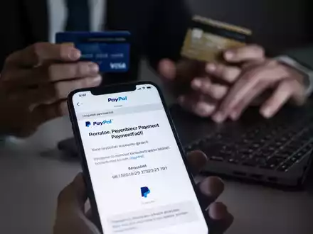 Ein Smartphone-Bildschirm zeigt eine betrügerische PayPal-E-Mail mit einer unmöglichen Frist. Im Hintergrund sind unscharf Hände und eine Kreditkarte zu sehen. - Foto: über boerse-global.de