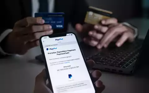 Ein Smartphone-Bildschirm zeigt eine betrügerische PayPal-E-Mail mit einer unmöglichen Frist. Im Hintergrund sind unscharf Hände und eine Kreditkarte zu sehen. - Foto: über boerse-global.de
