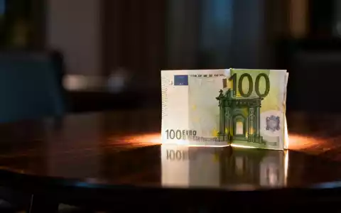 Eine gefaltete 100-Euro-Banknote liegt auf einem dunklen Holztisch mit weichem Licht von hinten. - Foto: über boerse-global.de