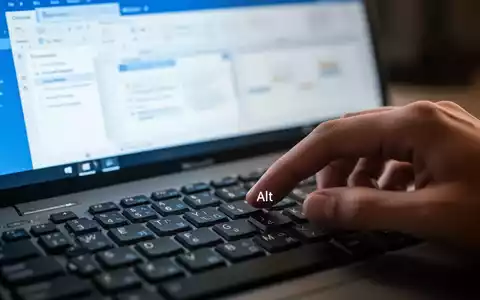 Nahaufnahme einer Hand, die die 'Alt'-Taste auf einer Computertastatur berührt, mit unscharfem Outlook-Bildschirm im Hintergrund. - Foto: über boerse-global.de