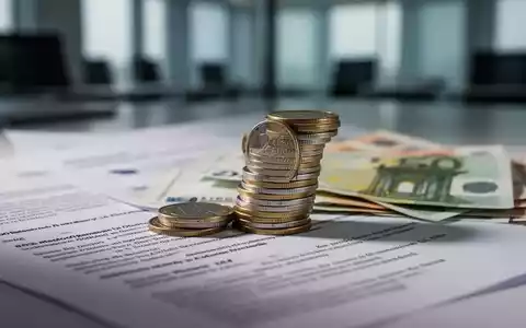 Ein Stapel Euro-Münzen und -Banknoten, teilweise verdeckt von Dokumenten, symbolisiert Sozialstaatsreformen und Sparmaßnahmen in Deutschland. - Foto: über boerse-global.de