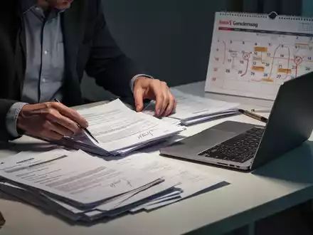 Eine Person sortiert Dokumente und einen Laptop auf einem Schreibtisch, mit einem Kalender im Hintergrund, der Termine anzeigt. - Foto: über boerse-global.de