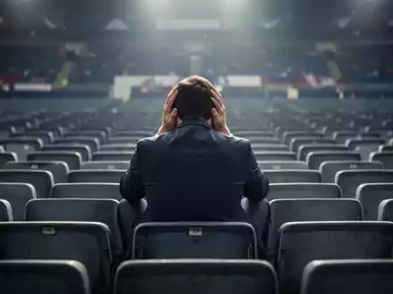 Ein einsamer Fußballfan sitzt enttäuscht mit dem Kopf in den Händen auf einem Stadionsitz, umgeben von leeren Reihen. - Foto: über boerse-global.de