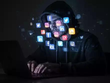 Eine Person im Kapuzenpullover tippt auf einem Laptop, mit leuchtenden Social-Media-Symbolen, die auf ihr Gesicht projiziert werden. - Foto: über boerse-global.de
