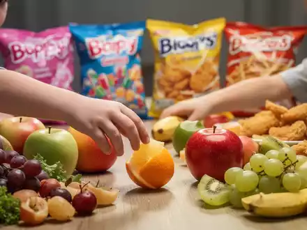 Eine Kinderhand greift nach Obst auf einem Tisch mit gesunden Lebensmitteln, im Hintergrund unscharfe ungesunde Snacks. - Foto: über boerse-global.de