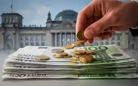 Eine Hand legt kleine Münzen auf einen Stapel Euro-Banknoten, mit dem Bundestag im unscharfen Hintergrund, symbolisiert Steuerreform. - Foto: über boerse-global.de