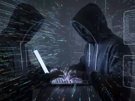 Verschattete Gestalt in Kapuzenjacke tippt auf Laptop, umgeben von digitalem Code und schwachen Blaupausen von Technikkomponenten. - Foto: über boerse-global.de