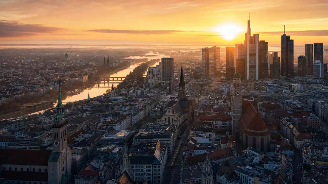 Dramatische Vogelperspektive der Münchner Innenstadt im goldenen Morgenlicht. Bürotürme und Kirchtürme zeichnen sich vor dem orangefarbenen Horizont ab. Rückversicherungs-Motiv für Munich Re, ISIN DE0008430026