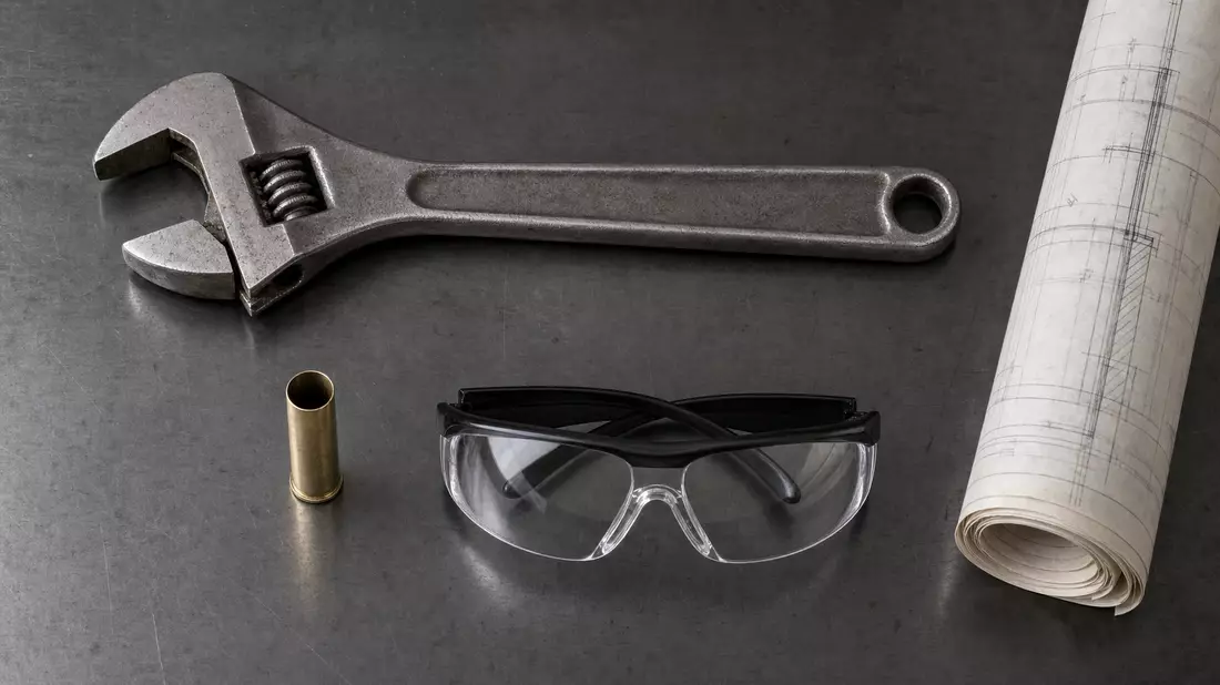 Flatlay mit Schraubenschlüssel, leerer Hülse, Schutzbrille und technischem Bauplan auf Holz