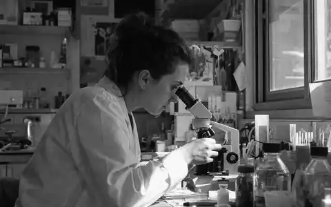 Qiagen N.V. (NL0012169213) Forscherin am Mikroskop, dokumentarische Schwarzweiß-Reportage mit körniger Textur, konzentriert - Foto: THN