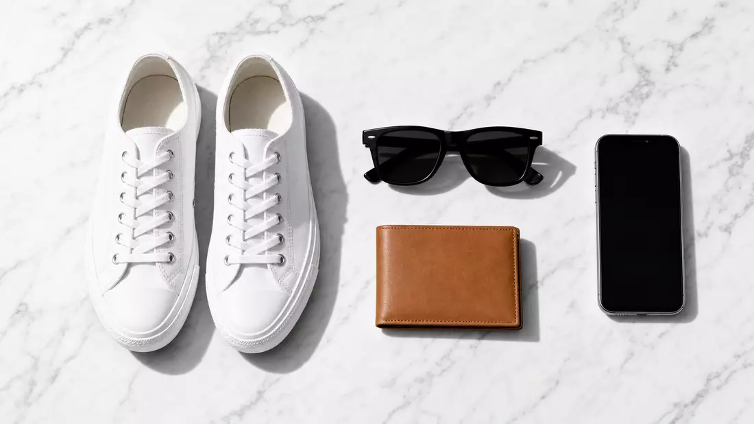 Flatlay mit weißen Sneakers, Sonnenbrille, Ledergeldbörse und Smartphone auf Marmor