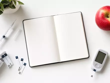 Novo Nordisk A/S (DK0062498333): Flatlay mit Spritze, Blutzucker-Teststreifen, Notizbuch und Apfel auf Weiß - Foto: THN