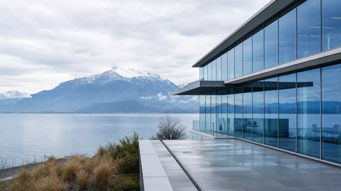 Modernes Glasgebäude am Genfersee mit Alpen im Hintergrund, bewölkter Himmel
