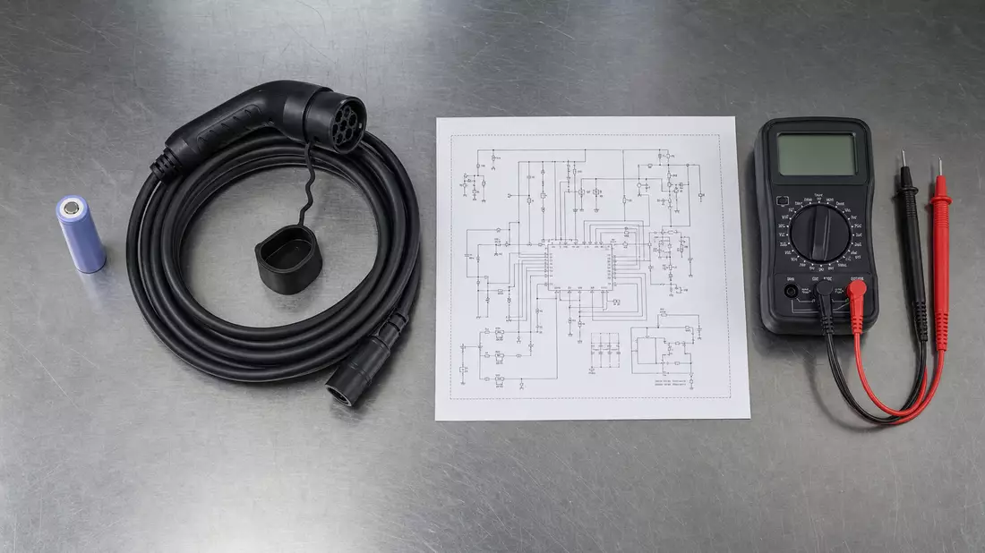 Flatlay mit Batteriezelle, Ladekabel, Multimeter und Schaltplan auf Edelstahltisch