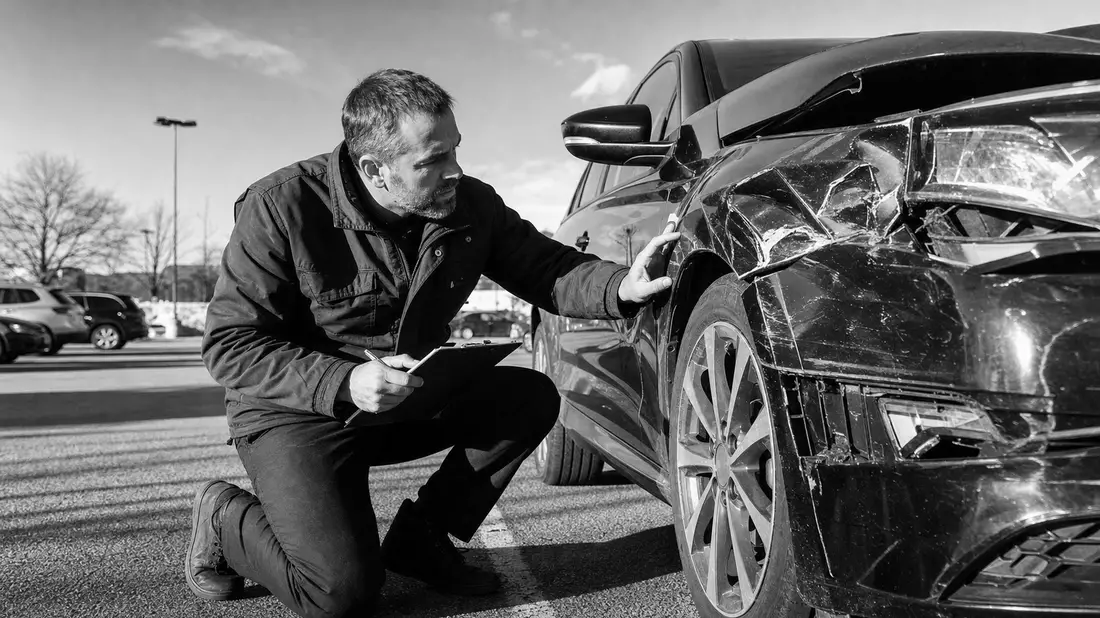Gutachter kniet neben beschädigtem Auto und notiert Schäden auf Klemmbrett