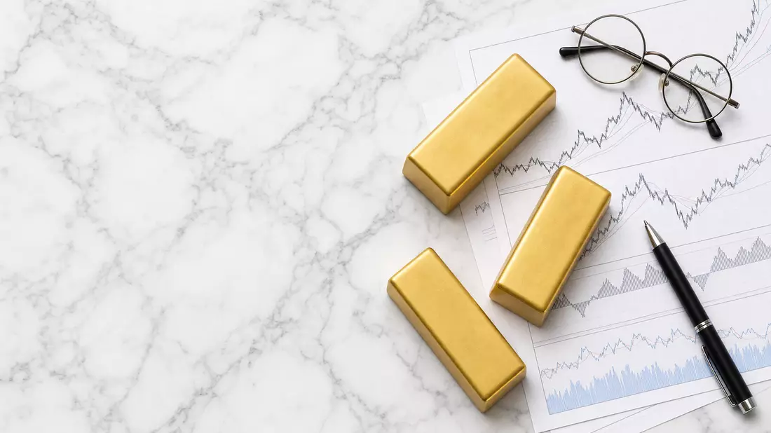 Goldbarren, Aktien-Charts, Brille und Stift auf Marmor-Oberfläche
