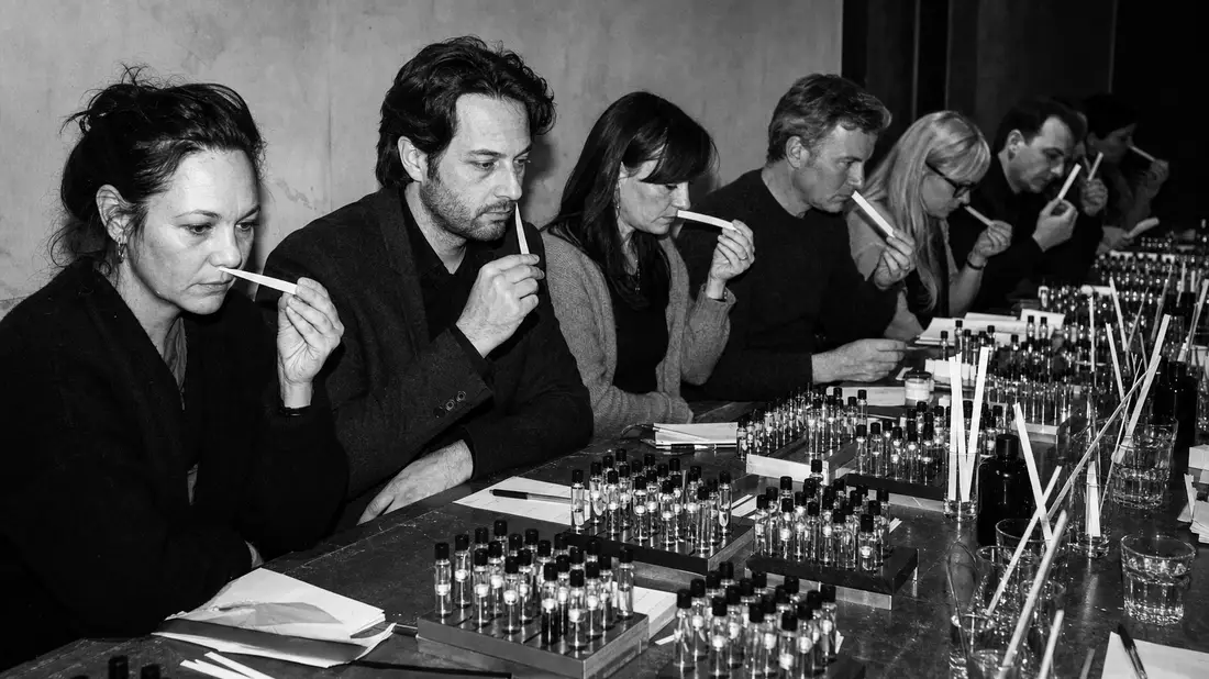 A high-contrast black and white documentary photograph of an aroma evaluation session. A row of tasters sit along a long table, each holding white scent strips to their noses. Rows of small unlabelled vials are arranged before them. Harsh directional flas