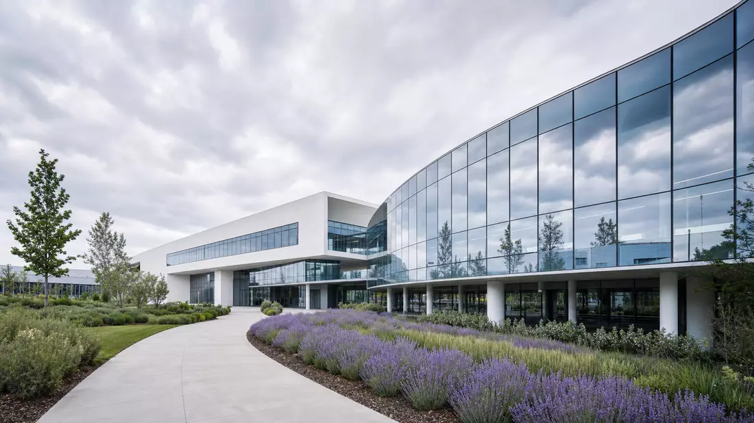 Architectural photography of a modern research and development building. A sweeping glass-curtain-wall wing reflects an overcast sky. A curved stone pathway winds through a landscaped garden with flowering lavender, rosemary, and aromatic shrubs. White co