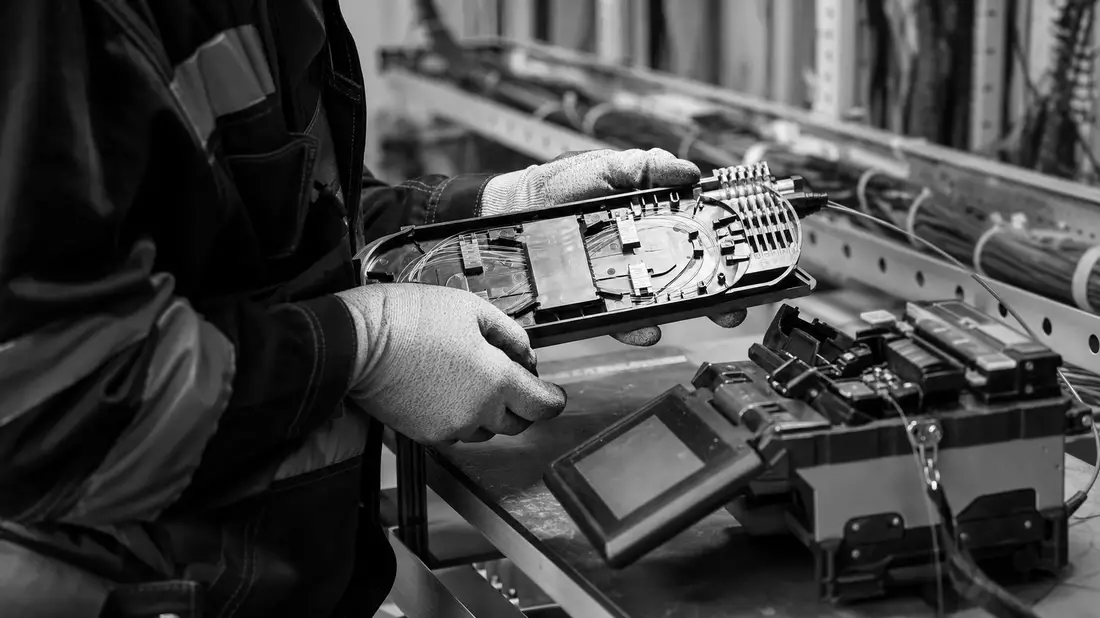 Black and white documentary photograph of a fiber optic technician in work gloves handling a fiber s