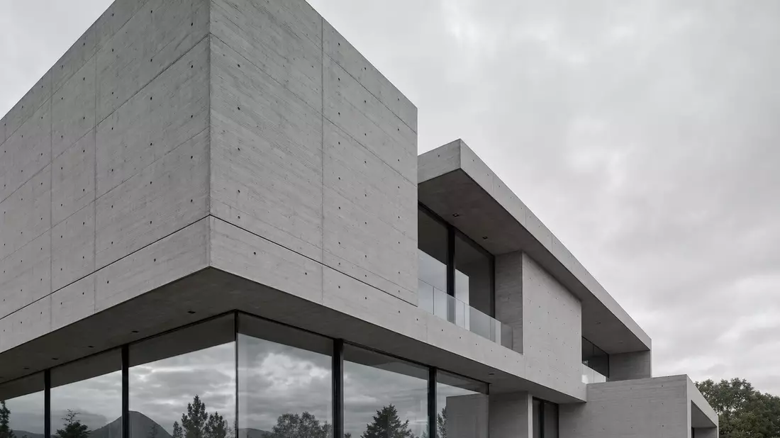 Architektonische Fotografie eines modernen Gebäudes mit expressiver Sichtbetonfassade. Präzise horizontale und vertikale Schalbrettfugen strukturieren die graue Betonoberfläche. Raumhohe Verglasung spiegelt den bewölkten Himmel. Strenge rechte Winkel und 