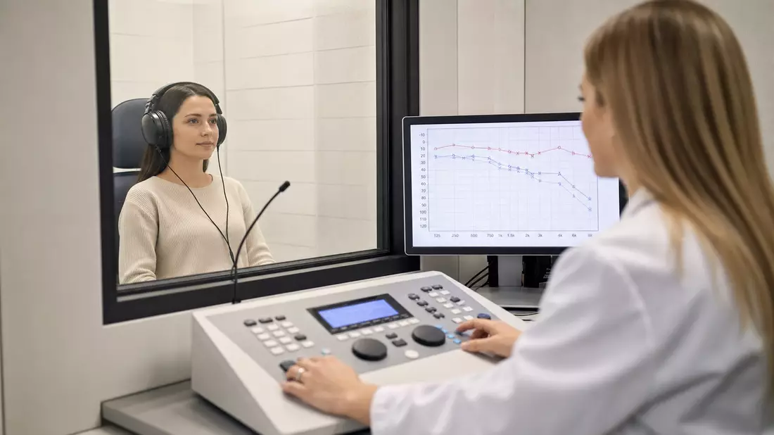 A photorealistic audiology examination scene showing a patient wearing large professional audiometry headphones seated in a sound booth, while an audiologist in a white coat operates a clinical control panel with an audiogram chart displayed on a monitor 