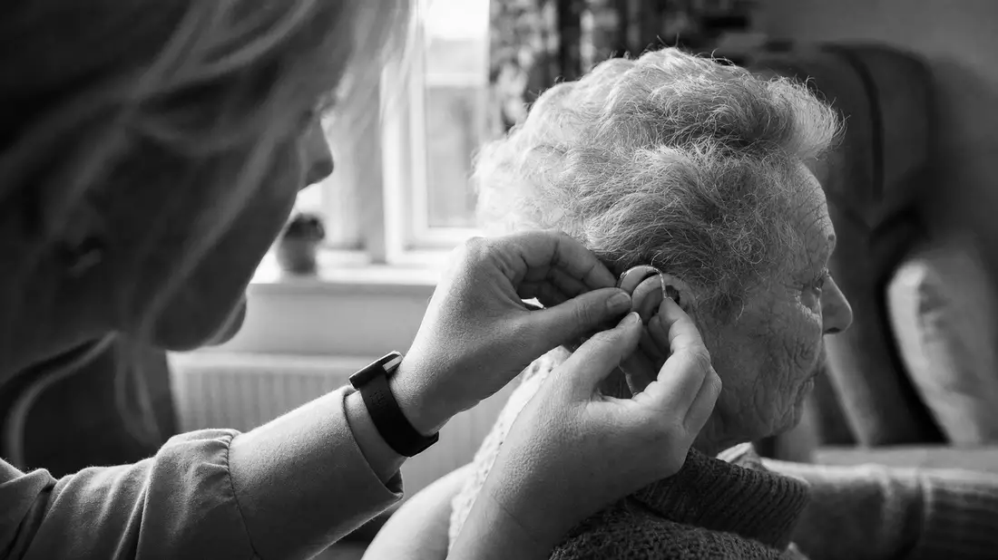 A black and white documentary reportage photograph showing a hearing care specialist's hands carefully fitting a small hearing aid behind the ear of an elderly patient with grey hair, captured in high contrast monochrome with natural window light and grai