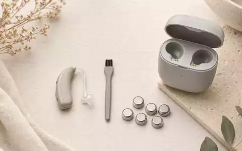 Sonova CH0012549785: Flatlay mit neutralem Hörgerät, Reinigungsbürste, Batterien und Ladestation auf Leinenstoff - Foto: THN