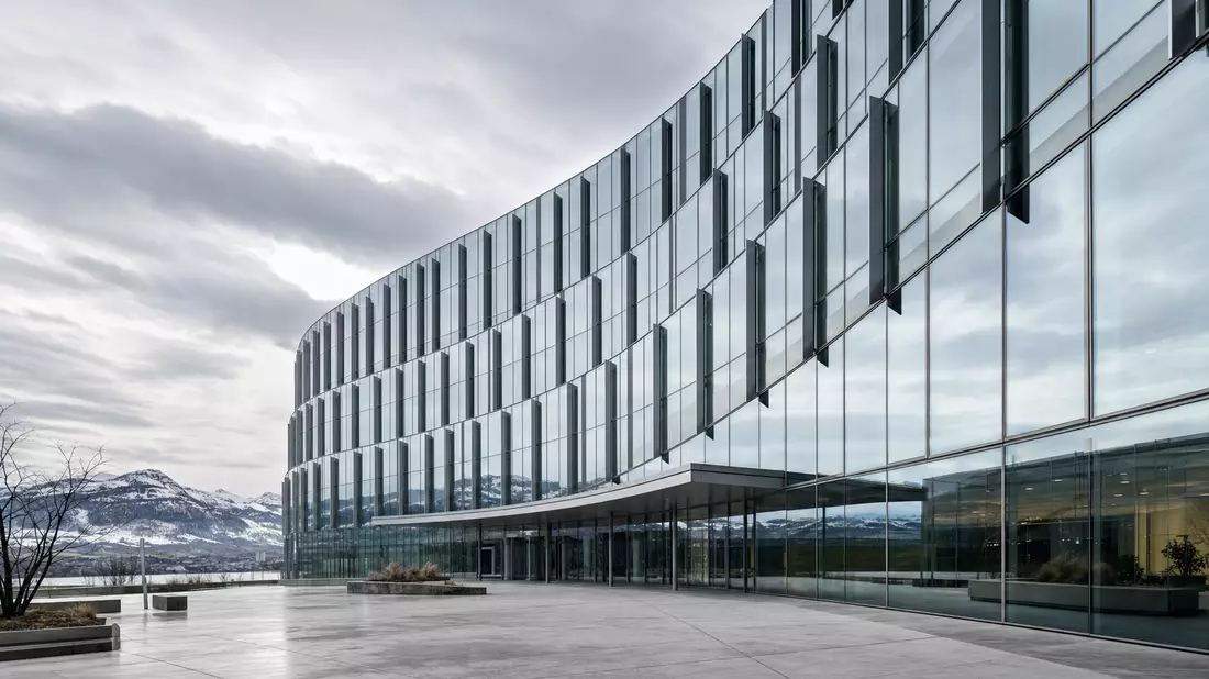 Architectural photograph of a contemporary corporate headquarters building in Zurich. A sweeping curved glass curtain-wall facade reflects clouds and the alpine skyline. Structural steel fins cast rhythmic shadows under an overcast northern light