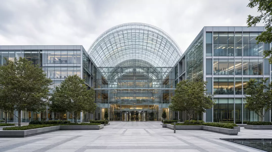 Architectural photograph of a prestigious financial services headquarters, central glass dome arching over a light-filled atrium visible from the exterior plaza, symmetrical curtain-wall facades in grey and glass, mature trees lining the paved forecourt, 