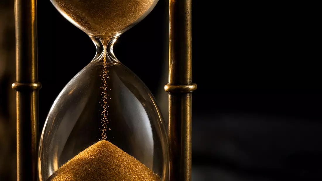 Extreme macro close-up of a brass-framed hourglass, fine golden sand flowing in a thin stream through the glass waist, sharp focus on individual grains mid-fall, dramatic raking sidelight from the left, deep shadow on the right, warm amber tones against n