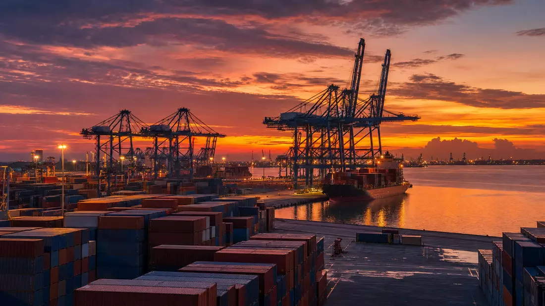 Wide panoramic photorealistic container port terminal at dusk, gantry cranes silhouetted against orange and purple sky, stacked shipping containers, calm water reflections