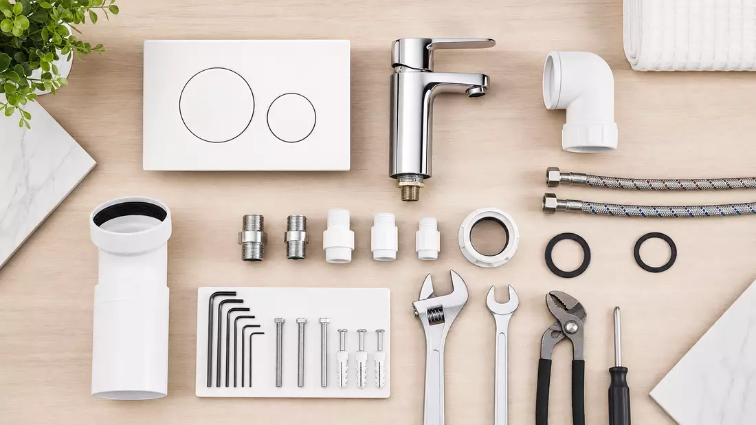 Top-down flatlay of sanitary installation components neatly arranged on light wood surface including white flush plate, chrome faucet, pipe connectors, wrenches, allen keys and rubber seals