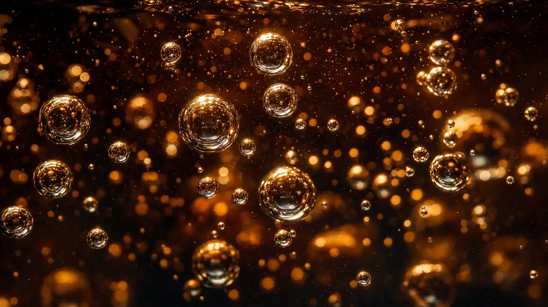 Extreme macro close-up of carbonation bubbles rising through dark caramel cola liquid, dozens of spherical transparent bubbles with bright backlit halos and light reflections, shallow depth of field with sharp foreground and dreamy background, dramatic hi