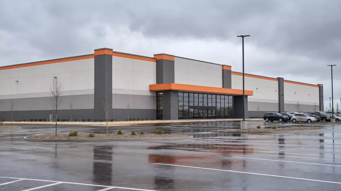 Architectural render of a large big-box retail warehouse building with subtle orange accent trim on facade, massive flat-roofed structure with parking lot in foreground, wide-angle view under overcast skies, no logos or signage