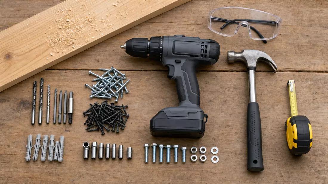 Overhead flat lay of home improvement tools neatly arranged on a wooden workbench: unbranded cordless drill, claw hammer, measuring tape, assorted screws, safety goggles, and a wood plank with sawdust under neutral daylight