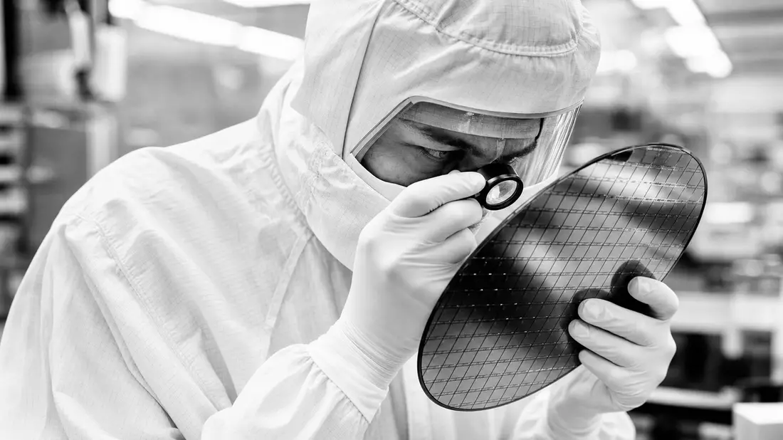 Black and white documentary photograph of a semiconductor engineer in a cleanroom bunny suit closely inspecting a silicon wafer with a jeweler loupe, intense focused expression, high contrast monochrome tones