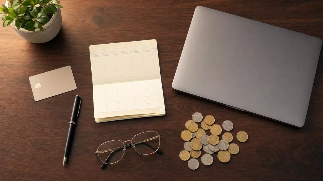 Overhead flatlay of banking essentials on dark mahogany: blank bank card, open savings passbook, black pen, wire glasses, scattered gold and silver coins, and closed laptop. Warm studio light, clean arrangement