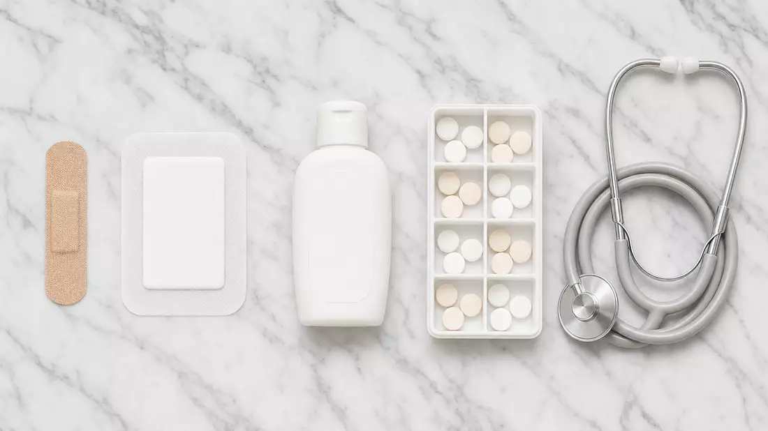 Overhead flat lay on white Carrara marble of five healthcare items in a neat row: a beige adhesive bandage strip, a white sterile wound dressing pad, a plain white lotion bottle, a white pill organizer tray with round tablets, and a coiled silver stethosc