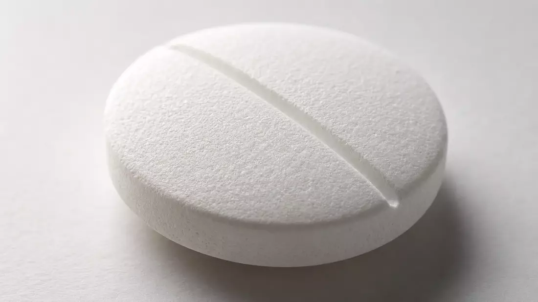Extreme macro photograph of a single white pharmaceutical tablet on a pale grey surface, a fine score line running diagonally across its center in sharp focus, micro-granular chalky texture visible across the pill surface, soft diffuse highlights along th