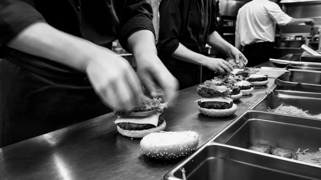 Schwarz-Weiß-Dokumentarfoto von Köchen beim Burger-Belegen in offener Restaurantküche, Edelstahl-Arbeitsflächen, Bewegungsunschärfe an Händen, dramatischer Kontrast, journalistischer Stil, kein Branding