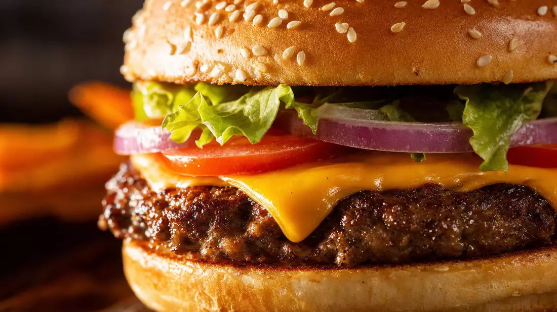 Makro-Nahaufnahme eines frisch belegten Burgers, dramatisches Seitenlicht, goldenes Sesambrötchen, schmelzender Käse, Salat- und Tomatenlagen, geringe Tiefenschärfe, warme Töne, kein Branding