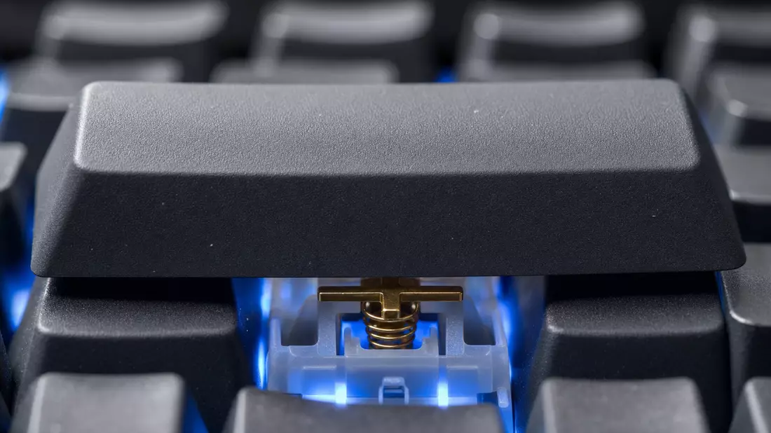 Extreme macro photograph of a single wide mechanical keyboard keycap partially removed, revealing gold spring and translucent switch housing with blue backlight glow, surrounding keys in soft bokeh, high contrast studio lighting
