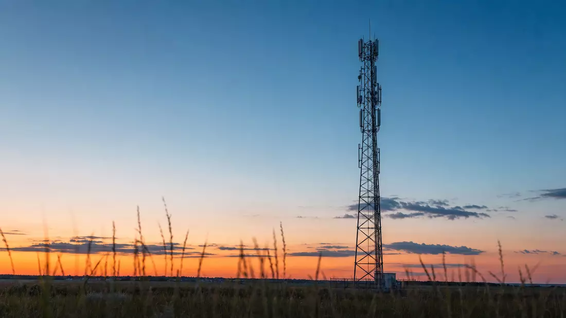 Ein schlanker 5G-Antennenmast aus Stahl steht bei Sonnenuntergang auf einem weiten Feld, orangeroter Abendhimmel, markenneutral
