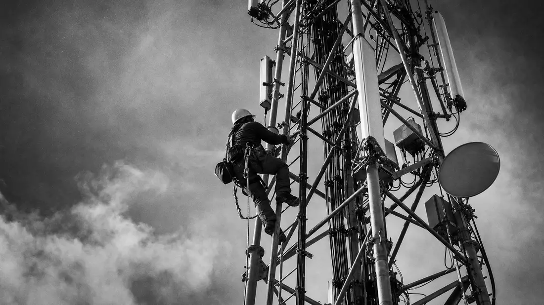 Schwarzweißes Reportagefoto eines Telekommunikationstechnikers mit Schutzhelm beim Klettern an einem Antennenturm vor dramatischem Wolkenhimmel