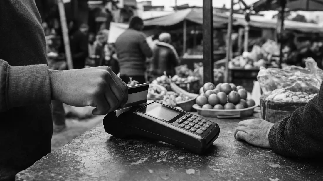 Schwarz-Weiß-Reportagefoto eines generischen POS-Terminals auf einem belebten Straßenmarkt – zeigt a