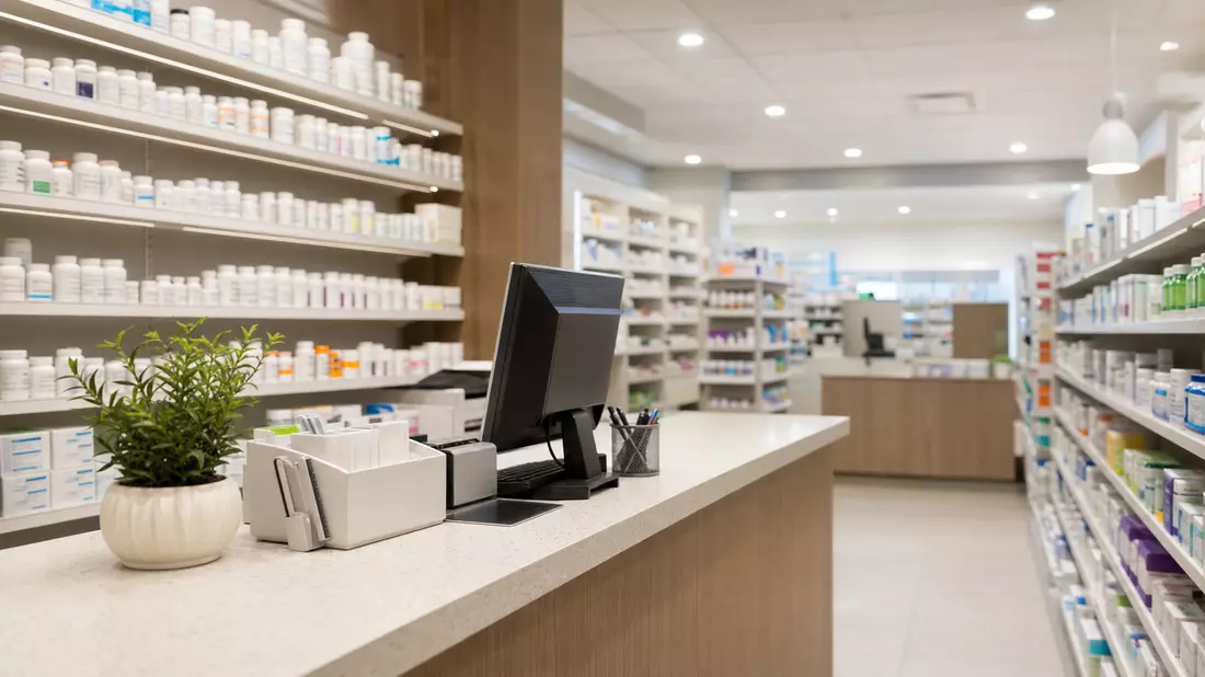 Helles, modernes Apothekeninterieur mit vollbesetzten Arzneimittelregalen und einem Rezeptpult im Vordergrund bei warmer klinischer Beleuchtung