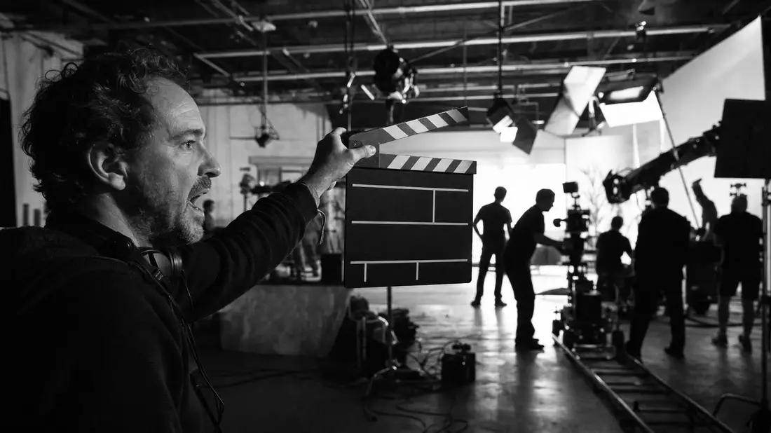 Schwarz-Weiß-Reportagefoto eines Filmstudios: ein Regisseur hält eine leere Klappe hoch, Crew-Silhouetten und Kameraequipment im kontrastreichen Hintergrund